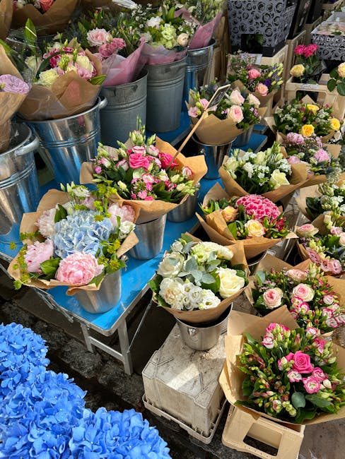 A display of freshly arranged floral bouquets and potted flowers at a flower market, featuring a variety of blooms including pink roses, white carnations, pastel hydrangeas, and light green foliage, a