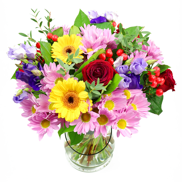 This mixed-colour bouquet, displayed in a simple clear glass vase against a white background, captures the warmth and bustle of everyday life in Acton. The composition is generous and rounded, with the eye naturally drawn to a cluster of deep red roses at the centre, their velvety petals folding in rich layers that speak of affection and timeless sentiment. Just ahead of them, bold yellow gerbera daisies stand proudly, each bloom framed by bright petals around a dark brown-orange centre, a combination that feels upbeat and welcoming, like stepping into a favourite café off Churchfield Road. Encircling these focal flowers is a ring of soft pink daisies or chrysanthemums, their many slender petals and dainty yellow centres forming a gentle, cushioning halo of colour that softens the arrangement. Threaded amongst the daisies and roses are slender, graceful stems of lisianthus in dusky purple and lilac, their bell-like blossoms adding a subtle elegance and a cool contrast that recalls late evening skies over Acton Green. Small clusters of round, glossy red hypericum berries peek out between the blooms, adding a playful, almost festive touch and a pleasing change of texture. Various greens in multiple shades-deep, sturdy leaves and light, wispy foliage-fill the spaces between flowers, giving the bouquet volume, balance, and a sense of natural movement, as though it were freshly gathered from a well-tended garden. Through the crystal-clear glass of the vase, neatly trimmed stems fan out into the water, revealing a professional, intentional design. Ideal for birthdays, get-well wishes, or simply brightening a home near Acton Central station, this bouquet feels both polished and heart-warmingly local.