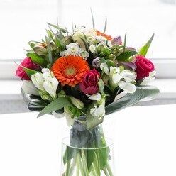 Masterfully arranged in a luminous setting that calls to mind Acton's sunlit terraces, this opulent bouquet balances colour, shape, and mood with floristic sophistication. Soft peach gerberas form a gentle frame, their creamy petals gracefully encircling brilliant orange centers that glint joyfully like lanterns on a summer evening in Acton Park. Regal magenta roses punctuate the display, their deep hue imparting richness and a feeling of romance, while airy sprays of ivory alstroemeria and fine white blossoms add layers of garden-inspired brightness. A verdant sweep of broad, glossy leaves-artfully curled as if crafted with care in a local boutique-grounds the exuberant flowers and brings harmony to the overall look. Pockets of smaller greenery and delicate sprigs slip between the florals, amplifying texture and dimension. Soft daylight spills across the bouquet, coaxing each colour into vivid relief without ever overpowering their subtlety. The clean fragrance-suggestive of lilies and dew-kissed greens-lingers, conjuring memories of leisurely walks past Acton's green squares. This arrangement's optimism makes it perfect for gatherings, celebratory brunches, or heartfelt tokens for anyone beginning a new journey within this dynamic London neighbourhood. Each bloom, each curve, is crafted with the attention and warmth that defines an expert Acton florist.
